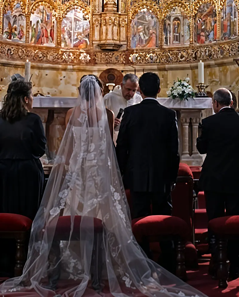 Ceremonia de boda religiosa en el altar de una iglesia, con los novios de espaldas frente al sacerdote y un impresionante retablo dorado con frescos religiosos al fondo.