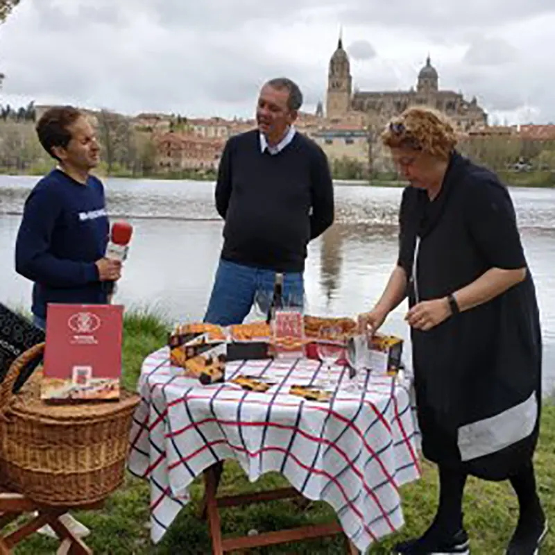 Grabación de una entrevista al aire libre junto al río Tormes, con la Catedral de Salamanca al fondo, una mesa con productos típicos y tres personas conversando en un ambiente distendido.