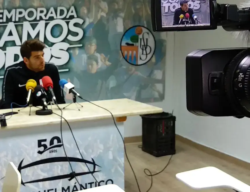 Rueda de prensa en el Estadio Helmántico con un jugador respondiendo a preguntas frente a varios micrófonos, mientras una cámara graba la escena. Al fondo, un cartel con el logotipo del club
