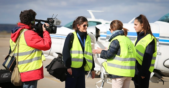 Equipo de grabación entrevistando a tres mujeres con chalecos reflectantes frente a una avioneta en un aeropuerto, en un entorno profesional vinculado a la aviación