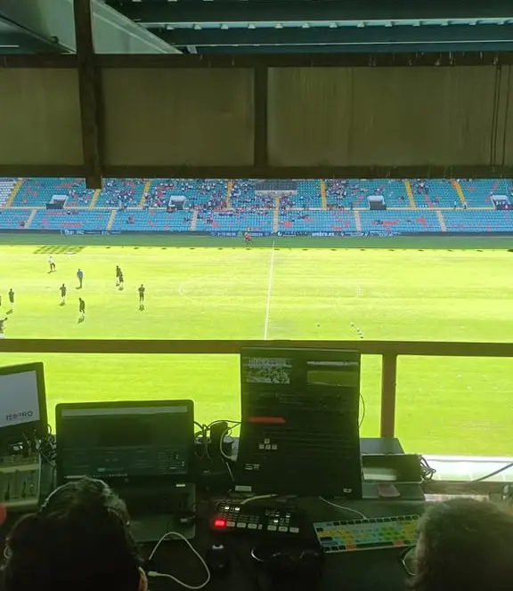 Puesto de realización audiovisual en un estadio de fútbol durante un partido, con monitores, mesa de mezclas y operadores controlando la retransmisión en directo desde la cabina de prensa.