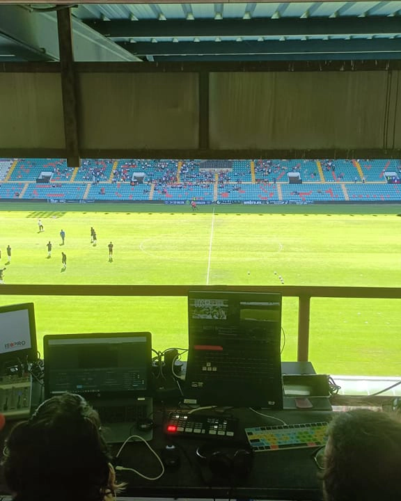 Puesto de realización audiovisual en un estadio de fútbol durante un partido, con monitores, mesa de mezclas y operadores controlando la retransmisión en directo desde la cabina de prensa.