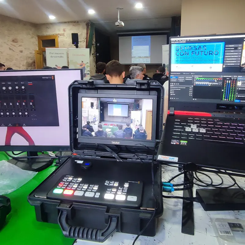 Equipo técnico para cobertura audiovisual de una jornada formativa con estudiantes, donde se ven cámaras, trípode y monitores de control en primer plano y un auditorio con asistentes al fondo. En pantalla "Dehesas con futuro"