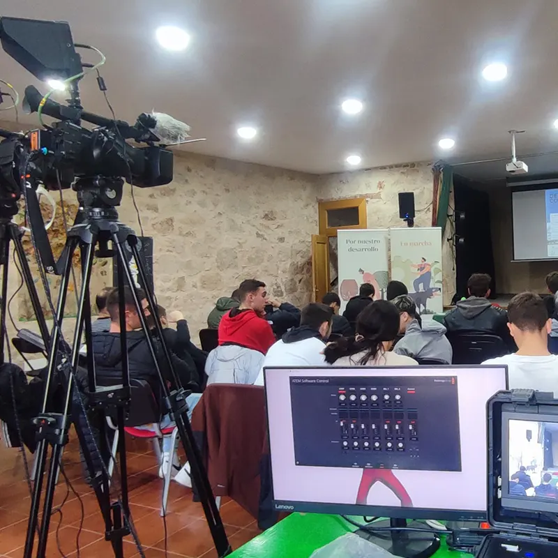 Cobertura audiovisual de una jornada formativa con estudiantes, donde se ve el equipo técnico con cámaras, trípode y monitores de control en primer plano y un auditorio con asistentes al fondo.