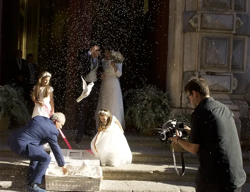 Salida de una boda con lanzamiento de arroz, palomas blancas y presencia de un videógrafo capturando el momento frente a una iglesia, mientras los novios celebran junto a niñas vestidas de blanco.