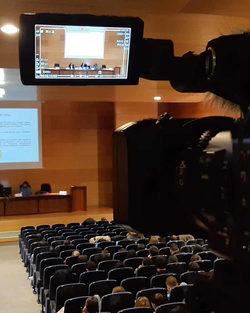 Grabación de una conferencia en un auditorio, capturada desde una cámara profesional que enfoca al escenario donde se desarrolla una ponencia con público asistente.
