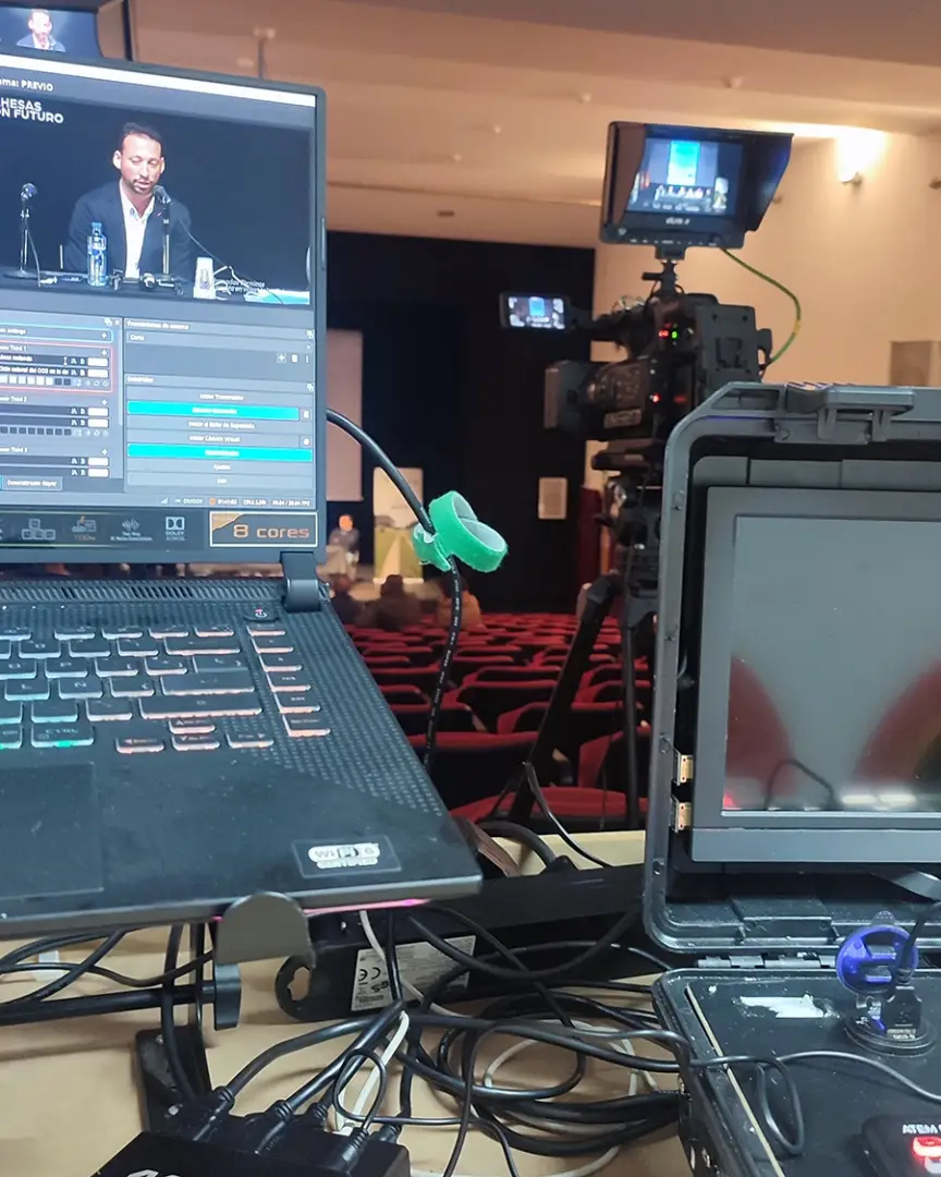 Equipo de producción audiovisual transmitiendo un evento en vivo, con ordenadores, cámaras profesionales y cableado en un teatro con público.