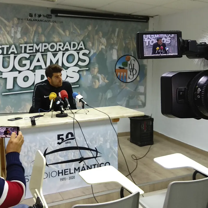 Rueda de prensa en el Estadio Helmántico con un jugador respondiendo a preguntas frente a varios micrófonos, mientras una cámara graba la escena. Al fondo, un cartel con el lema 'Esta temporada jugamos todos' y el logotipo del club.