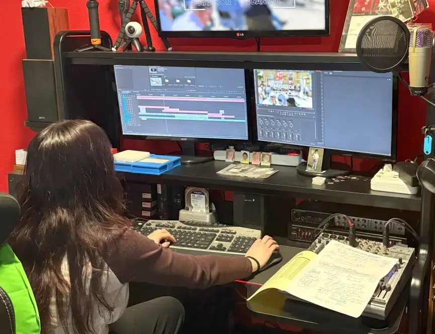 Editora de vídeo trabajando en un proyecto sobre el Museo de Automoción, con dos monitores, software de edición profesional y material técnico en una sala de postproducción con paredes rojas.